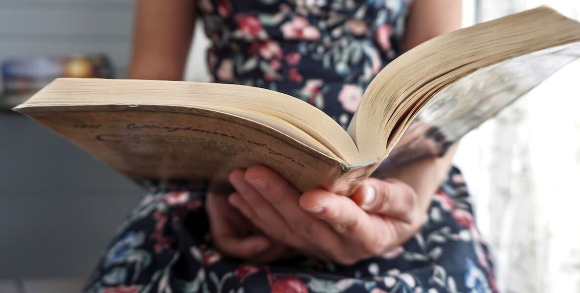 A woman wearing a black, floral-print dress sits near a window reading a book.