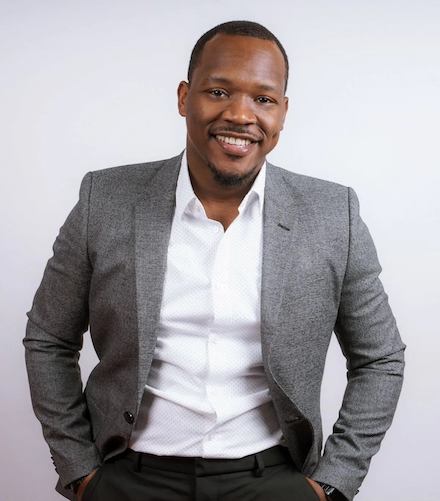 Raheem Manning, a Black man wearing a grey suit jacket and white button-down shirt has his hands in his pockets and smiles. He's Philly's "Night Mayor."