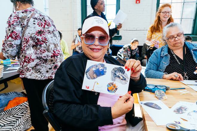 A woman wearing a pink baseball cap, large sunglasses, a dark shirt over a pink tee shirt smiles and holds her collage.