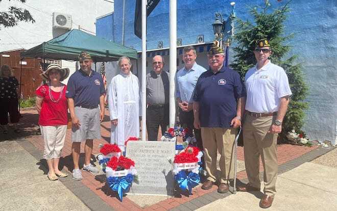 A group of 7 white people, including one priest in white, one woman in a red t-shirt, large hat and capris, and three men in army hats, stand on either side of a memorial plaque with red, white and blue flowers on either side in a small city park.