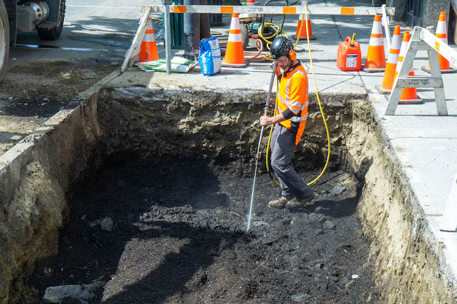 A worker in headphones, a helmet and neon shirt tends soil inside a tree well.
