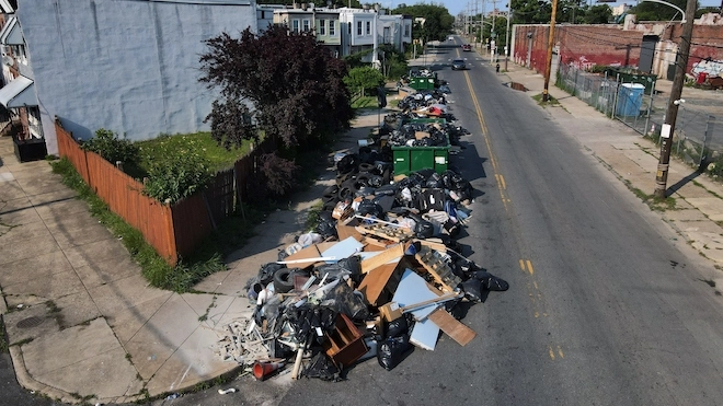 A giant pile of trash lines a Philadelphia street, overflowing out of green dumpsters.