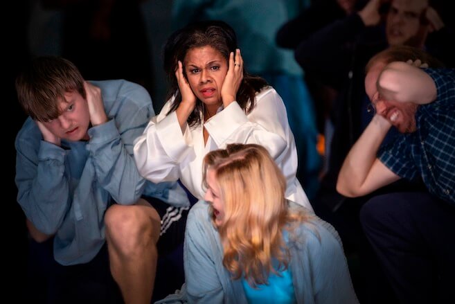 An African American woman in a white silk bathroom is surrounded by other people, all with hands on their ears in Opera Philadelphia's production of "The Listeners."