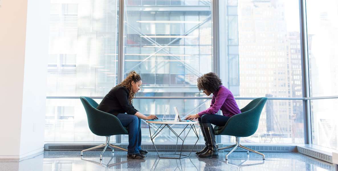 Two women type on laptops at a table in front of a giant window with an urban view