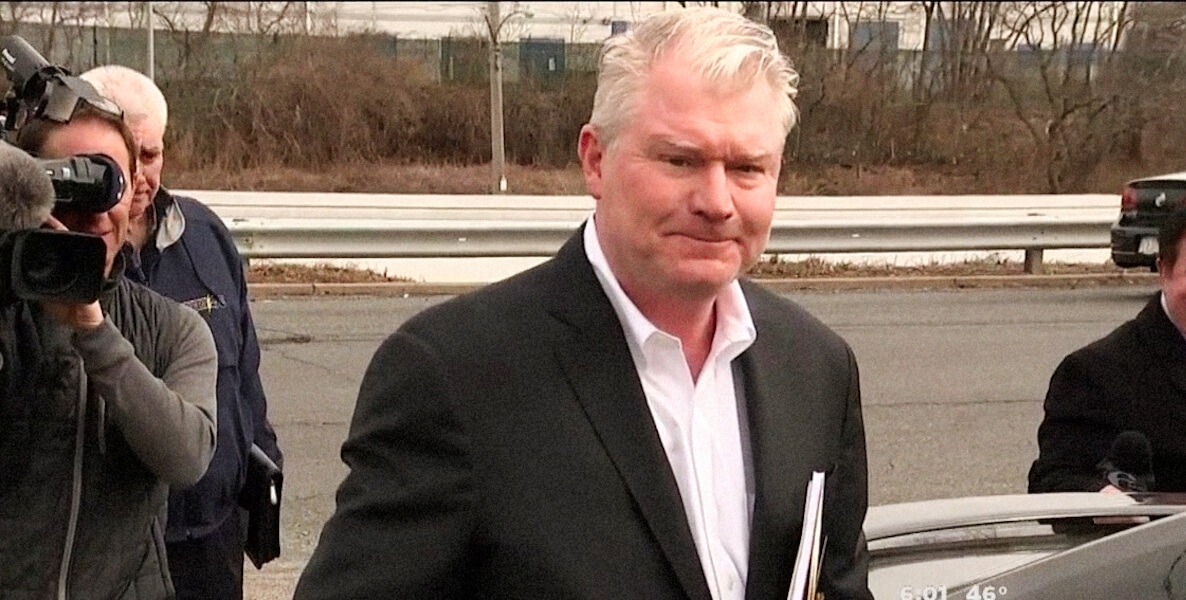 Philadelphia union boss John Dougherty has faced several corruption charges. Here, the White man with a pink face and sparse white hair wears a white button-down shirt with the top button open, a dark suit jacket, and carries documents under his left arm, with a member of the press pointing a TV camera at him. He appears to be in a parking lot.