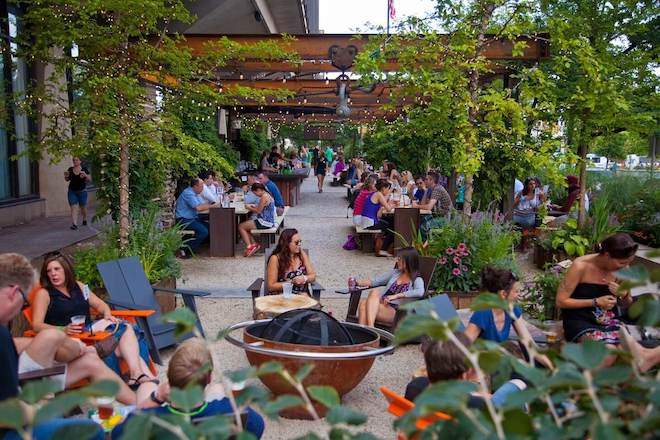 People sit in Adirondack chairs around an unlit firepit, among plants and trees. In the background, more people sit at covered area. The scene takes place next to what seems to be a large office building.