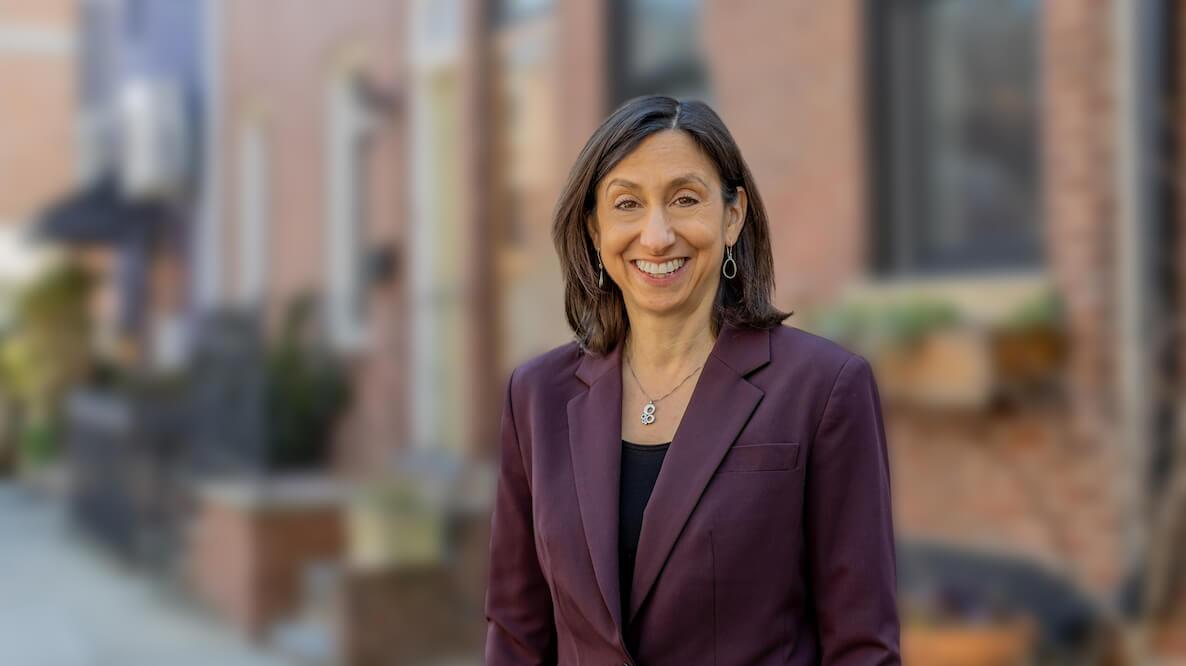Rue Landau, a civil rights and housing attorney and candidate for Philadelphia City Council, stands in front of a row of homes in Philadelphia. She is petite, with medium-length brown hair, wearing an aubergine jacket.