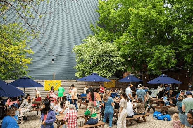 People sit and stand outside amid picnic tables on a gravel lot that's the temporary home to Mural City Wine Garden in Philadelphia.