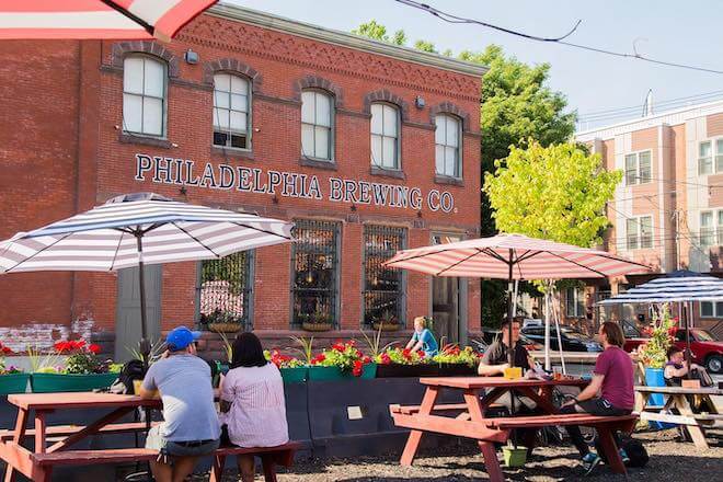 People sit at picnic tables with striped umbrellas outside Philadelphia Brewing Co., a brick brewery in Fishtown, Philadelphia