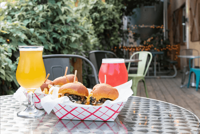Two poured beers flank small hamburgers (sliders) in a paper tray. behind them: Plants. At the Wissahickon Brewing