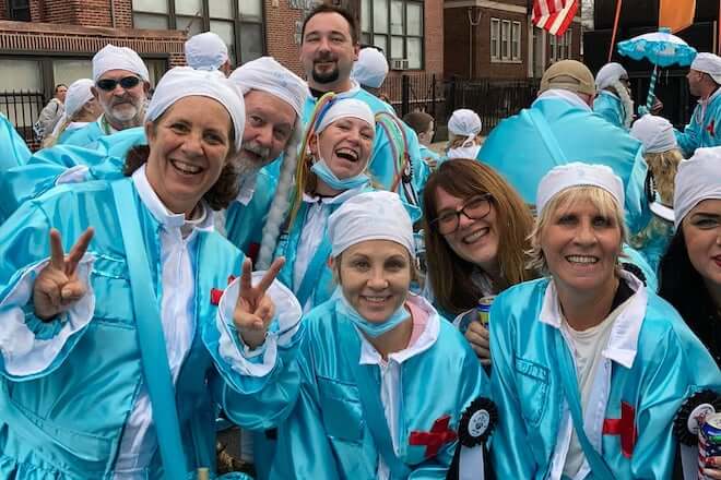 A crowd of White people wearing bright and shiny turquoise dresses and white caps stands outside, posing for the camera. They represent a comic club in the Philadelphia Mummers Parade.