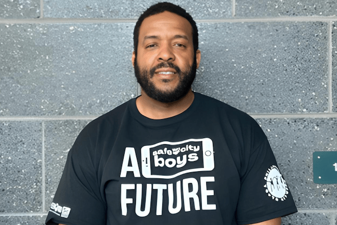 A Black man with a bear and mustache, wearing a black t-shirt with white writing stands in front of a cement block wall, smiling.