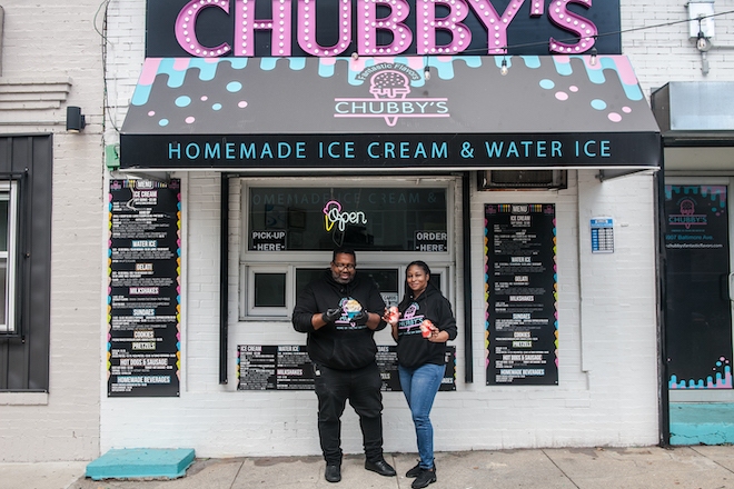 Tyson Devore, a Black man with a grey beard and glasses, wearing all black and Nikki Moore, a Black woman with long hair wearing a black Chubby's hoodie and jeans, stand outside Chubby's Fantastic Flavors at 59th and Baltimore. The couple both holds ice cream from their shop.