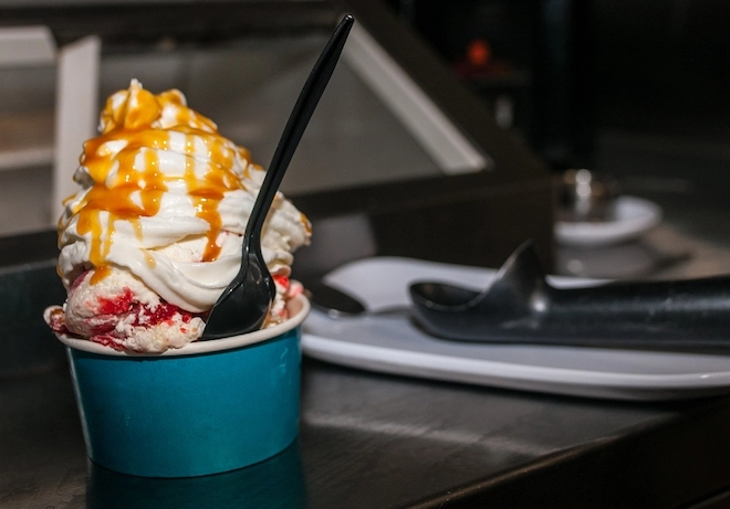 A blue paper cup holds strawberry shortcake ice cream topped with heavy whipped cream and caramel drizzle from Chubby's Fantastic Flavors. A plastic spoon sticks out of the side. Behind it is a metal ice cream scoop and glass ice cream case.
