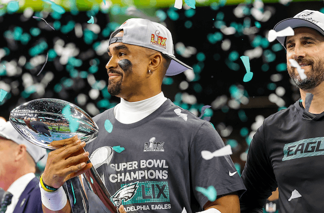 Jalen Hurts and Coach Nick Sirianni on the podium amid flying confetti. Hurts holds the Lombardi Trophy.