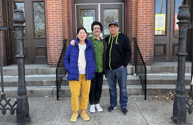 Artists Nadia Hironaka, an Asian woman in a blue puffer and yellow pants and shoes, Matthew Suib, a White man in a green shirt and dark pants, and participant Reverend Adan Mareina, a Latino man in a black cap and jakcet, jeans and sneakers, stand in front of a brick building.