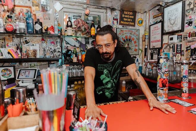 A white man with dark hair and beard and glasses stands behind a bright bar and in front of a sticker-covered wall at Tattooed Mom.