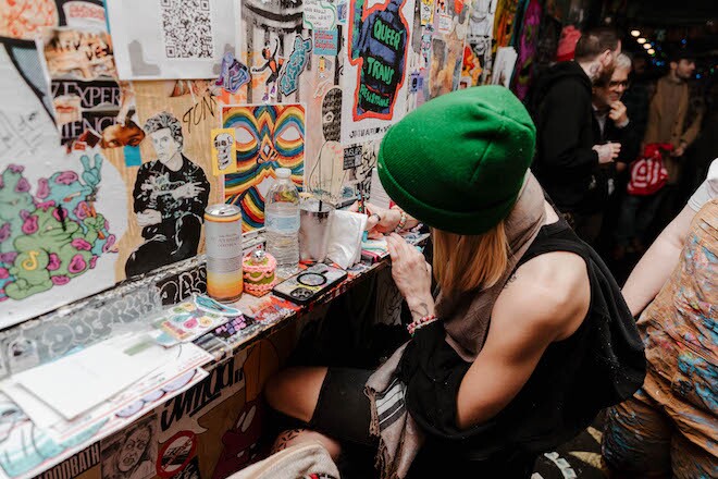 A white woman in a sleeveless top and green beanie works on a wheatpaste on a wall of Tattooed Mom.