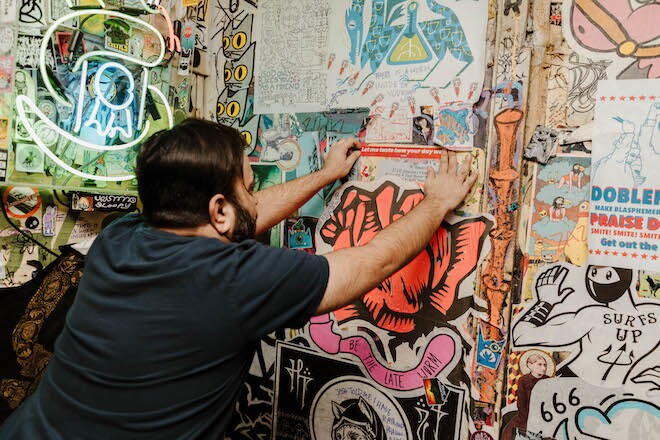 A white man with dark short hair and beard and dark t-shirt hangs a sticker on the wall of stickers at Tattooed Mom.