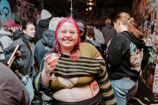 A white woman with pink hair, glasses, a string bikini top and cropped striped sweater holds a cocktail at a party upstairs at Tattooed Mom.