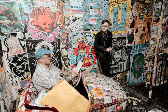 A white man in a sweater and cap sits in a former bumper car covered in stickers; another white man leans against one of two walls covered in wheatpastes and stickers.