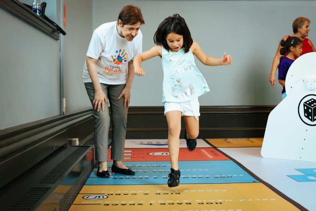 Kathy Hirsh-Pasek, an older white woman with short hair and a playful learning t-shirt, observes a child jumping on a floor map in Please Touch Museum's Parkopolis exhibit.