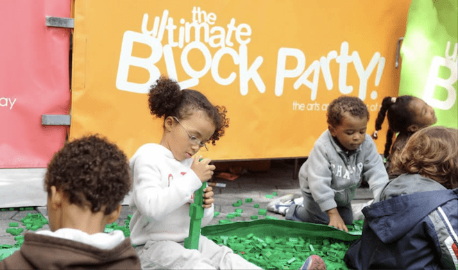 Small children sit on faux grass playing with toys in front of a sign that says "The Block Party."