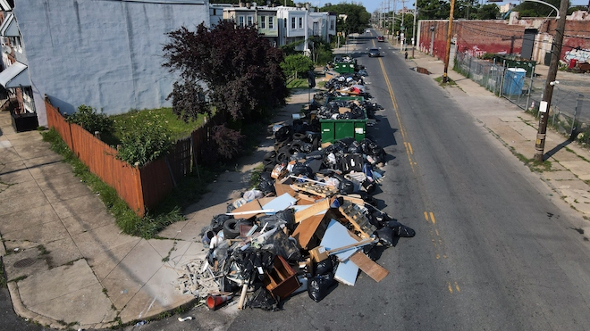 A giant pile of trash lines a Philadelphia street, overflowing out of green dumpsters.