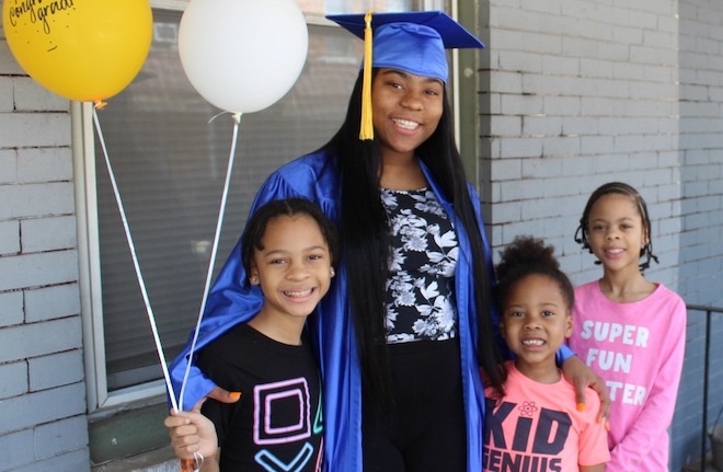 An African American woman in cap and gown holds balloons and puts her arms around three girls. She's a graduate of Beyond Literacy in Kensington Philadelphia.