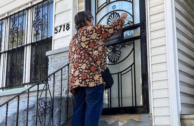 A person in a floral top and jeans knocks on the storm door of a home with aluminum siding. This person is a volunteer for Changing the Conversation Together, a deep canvassing group in West Philadelphia.