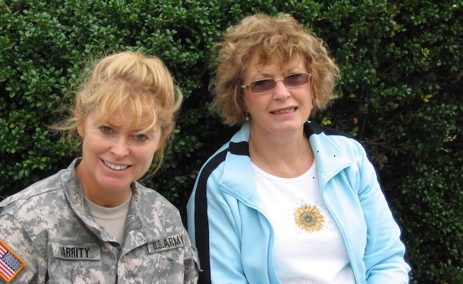 Pennsylvania State Treasurer Stacy Garrity, a white woman with fluffy strawberry blonde bangs long hair pulled back, wears U.S. Army fatigues and smiles alongside her mother, who has shorter wavy brown-blonde hair, small transitions glasses, a blue and black track jacket, unzipped, and a white shirt with a small sunflower in the center of it.