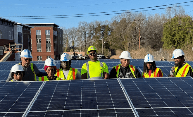 Eight Black Richmond, VA residents wearing hard hats and safety vests stands among solar panels outdoors.