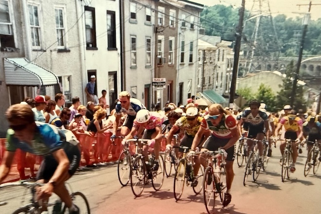 Cyclists on racing bikes ride up a steep hil past rowhomes in the Manayunk neighborhood of Philadelphia.