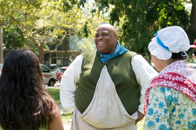 Michael Idriss, a tall bald Black man in colonial garb, stands outside in a park, facing costumed and non-costumed onlookers, and smiling.