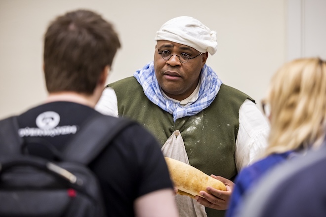 Michael Idriss, a tall Black man in colonial chef's garb and a wired spectacles, holds a loaf of bread while facing non-costumed onlookers.
