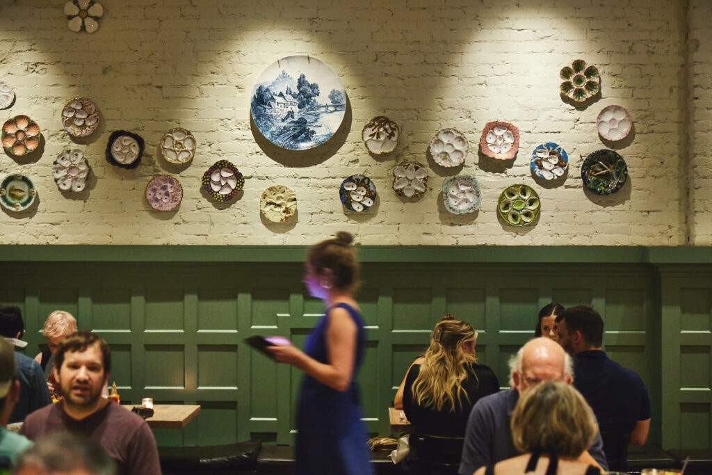 Diners sit at tables, some against a green panel wall. Overhead, antique and vintage oyster platters hanging on a white-painted brick wall, part of the Oyster House restaurant in Philadelphia.