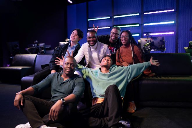 A group of young entrepreneurs posing together on a lounge seat at a neon-lit gaming expo. They are Left to right, back row: Rory Soh, Nicodemus Madehdou, Manyeuris Soriano, and Godavari Hughes. Seated: Calbert Warner and Michael Berthaud, all Jumpbutton staff.