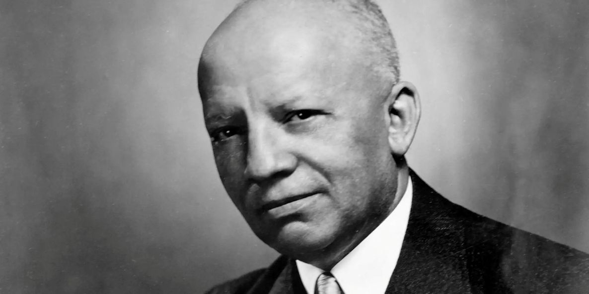 A black and white image of an American Black man named Carter Woodson. He is balding, wearing a dark suit and tie, and slightly smiling.