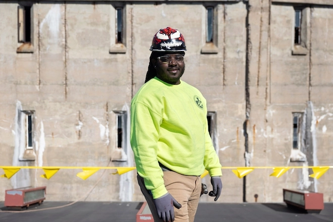 Construction academy student Mecchi Stewart-Rice wears a colorful hard hat, clear glasses, a bright yellow sweatshirt, grey gloves and khakis outside Eastern State Penitentiary.