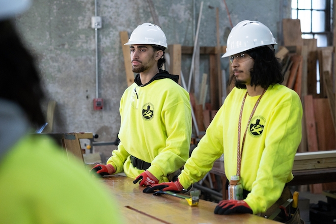 Two Latino students wear bright yellow sweatshirts, red gloves and white hard hats while standing at a construction work table.