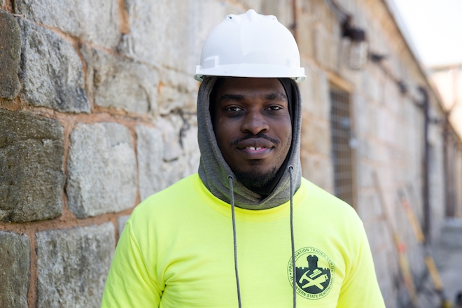 Carl Moisette, in a bright yellow sweatshirt, hoodie and white hard hat, smiles in front of a stone brick wall at Eastern State Penitentiary.