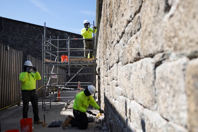 Three students in bright yellow sweatshirts, white hard hats and jeans works to replace mortar on a stone brick wall at Eastern State Penitentiary. One stands on scaffolding.