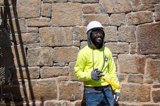 Abou Hinson, in a bright yellow sweatshirt, white hard hat, gloves and jeans, smiles in front of a stone brick wall at Eastern State Penitentiary.
