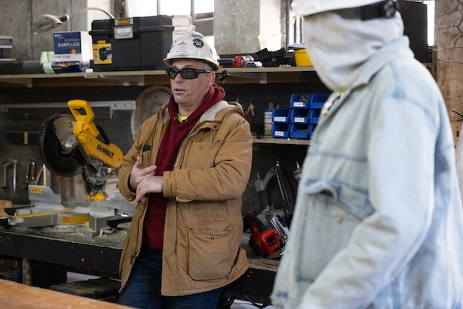Construction academy professor Chris Capaldi, a white man in a white hard hat, dark goggles and a work jacket, speaks to students (largely not pictured) among tools.