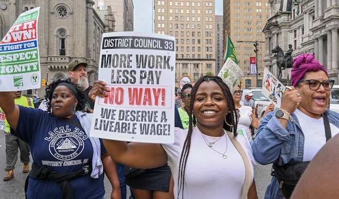 A Black woman repping DC 33 municipal workers holds a sign that says, "MORE WORK LESS PAY NO WAY!" outside Philadelphia City Hall.
