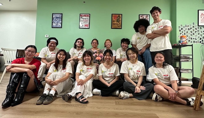 A group of high school and university students sits (and one stands) on a couch and the floor of the Ginger Arts Center. They are all wearing white t-shirts.