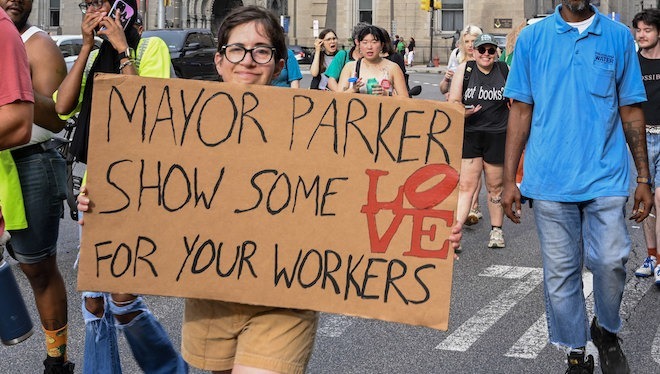A white woman with glasses supports striking DC 33 workers. She holds a cardboard sign that says: "MAYOR PARKER, SHOW SOME LOVE FOR YOUR WORKERS" outside Philadelphia City Hall. The "LOVE" resembles the Robert Indiana statue.