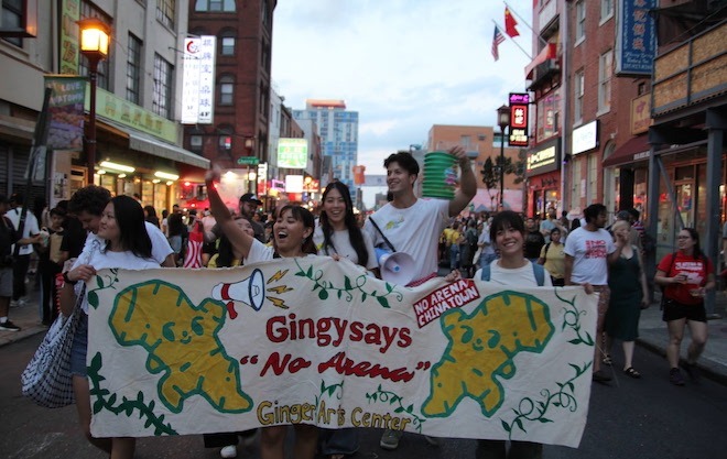 Asian students from the Ginger Arts Center carry a banner in Chinatown protesting the 76 Place arena. The sign says "Gingy says "No Arena" Ginger Arts Center" and depicts two smiling ginger root characters.