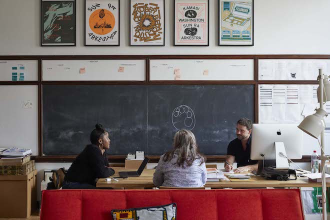 In Ars Nova studio, inside an old classroom with a blackboard, three people sit around a table talking.
