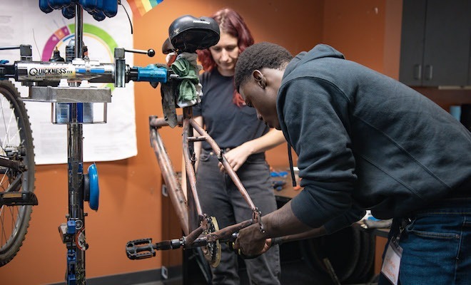 An African American student in jeans and a black hoodie leans to work on bike repairs to a frame mounted in a workshop. A teacher watches.